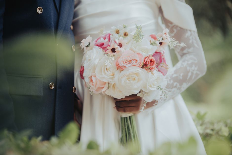 Close-up of a bride and groom holding a floral bouquet during a wedding. The bouquet features large, creamy white roses mixed with soft pink roses, small white daisies with dark centers, and delicate