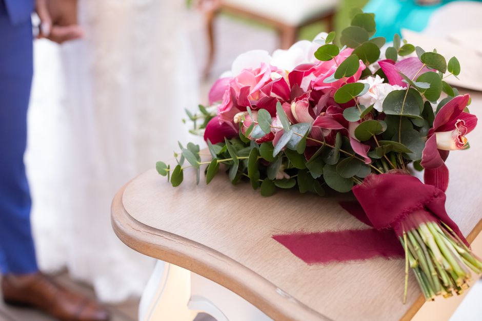 A bouquet of fresh flowers resting on a light wooden table, featuring pink and white roses, deep red calla lilies, and lush green eucalyptus leaves arranged in a compact, elegant style. The bouquet is