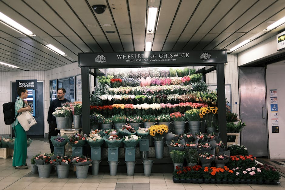 A floral display at Wheeler of Chiswick inside a subway station, featuring a variety of fresh flowers arranged in metal buckets and wrapped bouquets. The arrangement includes roses, tulips, daisies, a