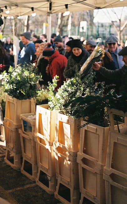 A floral market stall featuring fresh flower arrangements with lush greenery and vibrant blooms, displayed in beige wooden crates. The flowers include assorted foliage and flowering plants, arranged i