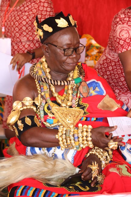 A woman dressed in traditional Maasai attire, wearing a vibrant red shuka with intricate beadwork and ornate gold jewelry, including necklaces, bracelets, and a headdress with black and gold accents. 